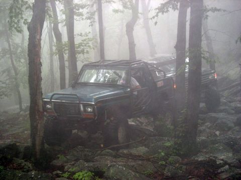 1988 Ford Bronco - A green doorless full-size 4x4 with a heavy brush guard, auxiliary lights and an exposed roll cage picks its way over wet boulders in dense…