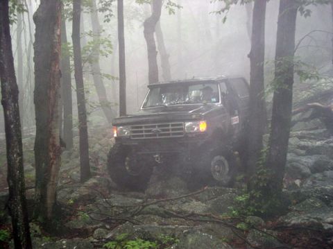 1988 Ford Bronco - A black Ford Bronco drives through a misty, wooded area with many rocks.