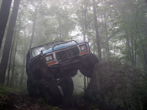 1988 Ford Bronco - Off-road SUV navigates a large rock obstacle in a misty wooded area.