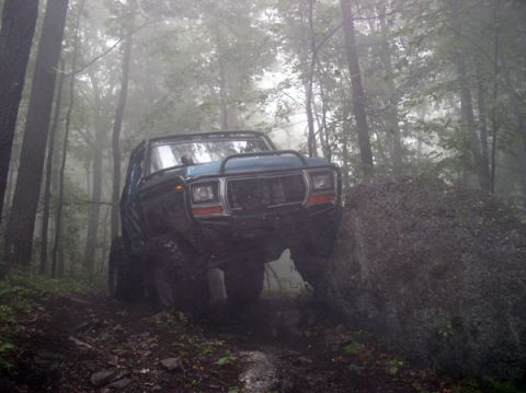 1988 Ford Bronco - A blue SUV navigates a challenging off-road trail through a misty wooded area.