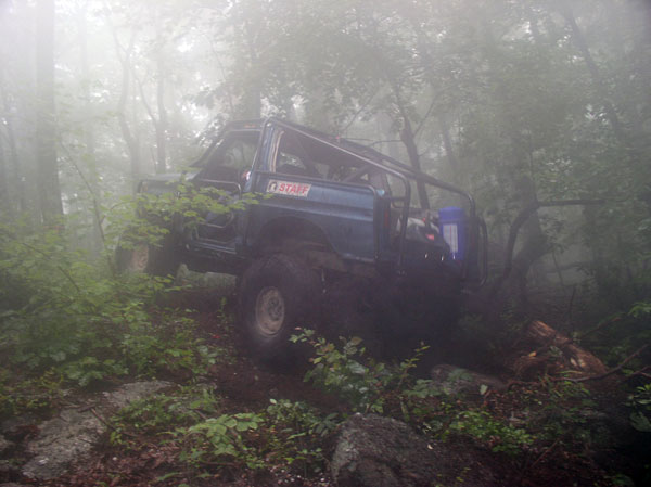 Blue topless caged 4x4 with mud tires flexing over roots on a misty forest trail