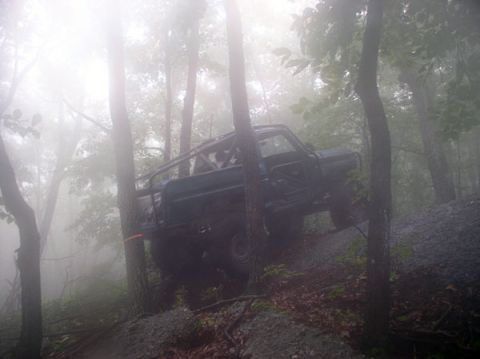 1988 Ford Bronco - A modified off-road vehicle tackles a steep, wooded incline shrouded in fog.