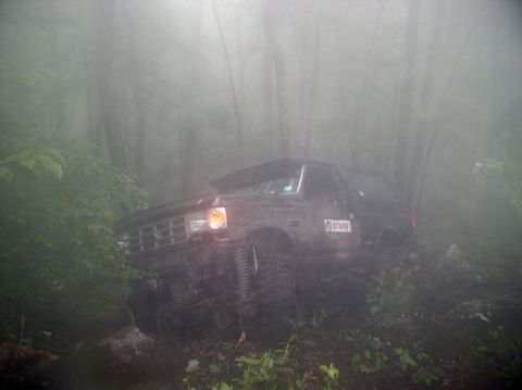1988 Ford Bronco - A dark-colored Ford Bronco navigates a wooded trail shrouded in fog, with a 'STAFF' sticker visible on its side.