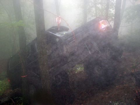 1988 Ford Bronco - A 1988 Ford Bronco is stuck between trees in a muddy, foggy forest scene, with its front headlight illuminated.