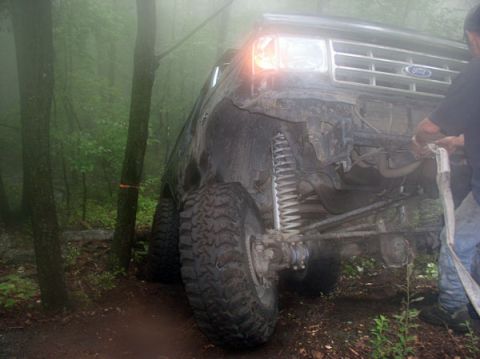 1988 Ford Bronco - Low front-corner view of a mud-caked Ford 4x4 on a foggy trail with the front bumper removed, exposing the coil spring, radius arm, solid a…