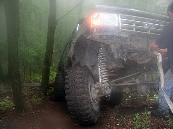 Muddy Ford 4x4 with exposed front coil spring and axle being strapped for recovery in fog