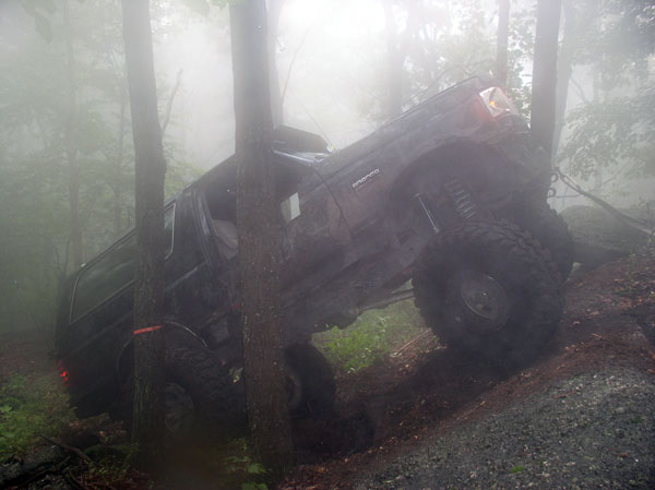 Black SUV tilted on a muddy incline, surrounded by trees and fog. Large off-road tires are visible
