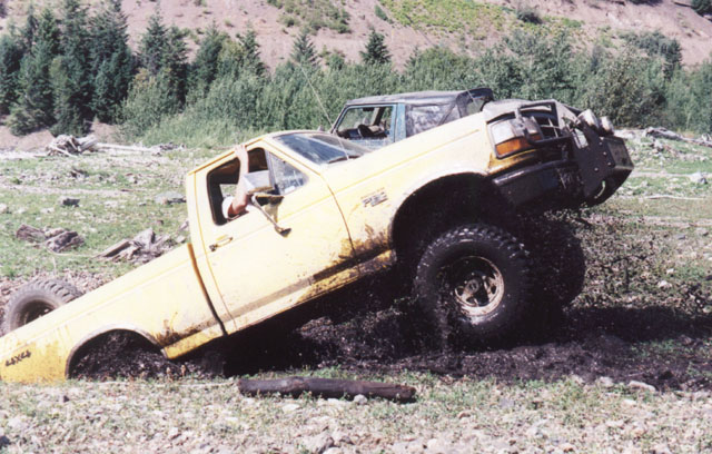 Muddy yellow Ford F-150 pickup nose-down and leaning in a mud pit
