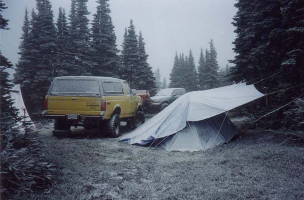 Lifted yellow Ford pickup with camper shell at a snowy pine campsite next to a canvas tent
