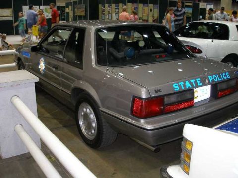 1998 Ford Crown Victoria - Rear three-quarter view of a silver/gray Ford Mustang LX Fox-body coupe marked 'STATE POLICE' in blue lettering on the trunk, displayed ind…