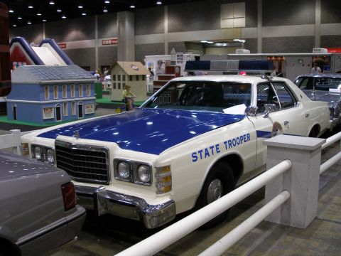 1998 Ford Crown Victoria - A white and blue vintage full-size sedan marked 'STATE TROOPER' with a blue roof-mounted light bar, displayed at an indoor show. The car is…