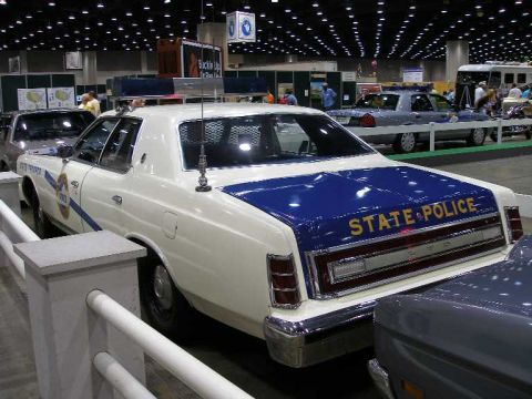 1998 Ford Crown Victoria - Rear three-quarter view of a restored late-1970s Ford LTD state police cruiser at an indoor car show, white with a blue hood and trunk lid…