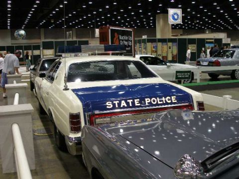 1998 Ford Crown Victoria - Rear three-quarter view of a white-over-blue 1970s Ford LTD state police cruiser displayed at an indoor car show, with STATE POLICE letteri…