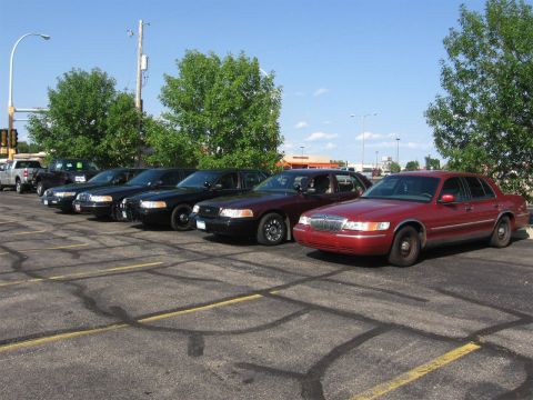 1999 Ford Crown Victoria - A line of sedans, including a red Mercury Grand Marquis, are parked in a lot on a sunny day.