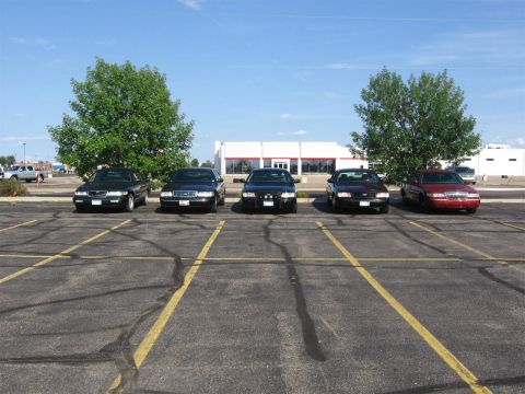 1999 Ford Crown Victoria - Multiple sedans are parked in a lot under a clear blue sky with a building and trees in the background.