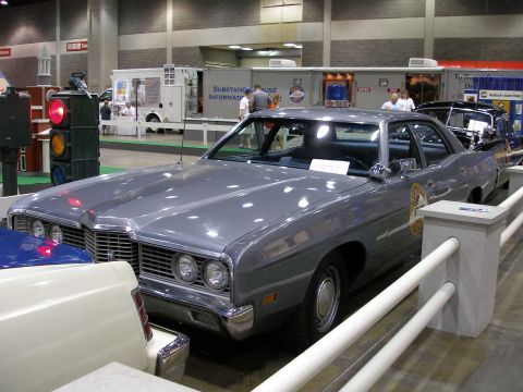 1998 Ford Crown Victoria - A gray full-size Ford four-door sedan displayed at an indoor show, shown from a front three-quarter angle with a round gold door emblem and…