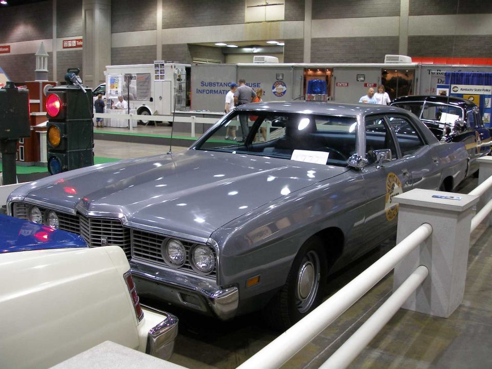Gray vintage Ford four-door sedan police car at an indoor show, 3/4 front view