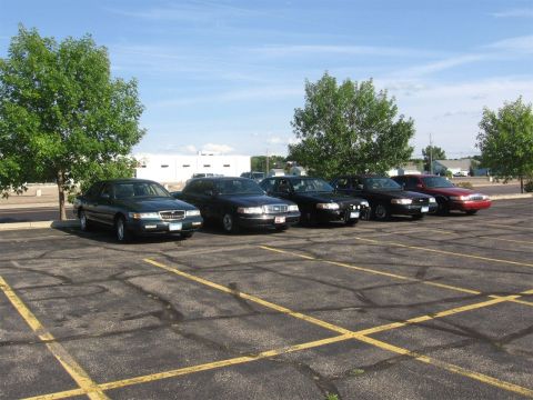 1999 Ford Crown Victoria - A row of sedans, including a 1999 Ford Crown Victoria, are parked in a lot under a blue sky.