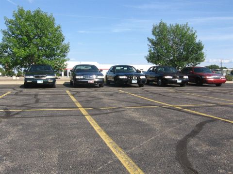 1999 Ford Crown Victoria - Several sedans are parked in a lot under a clear blue sky with trees and a building in the background.