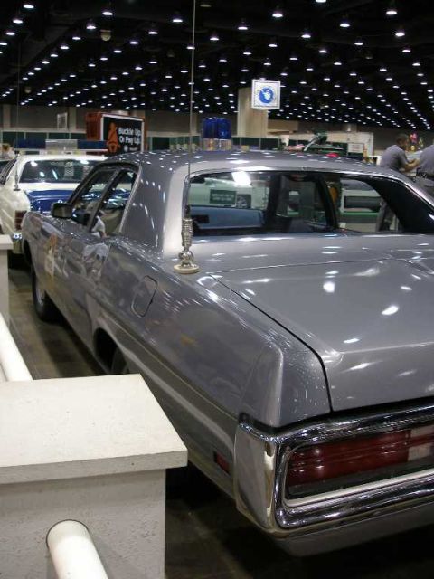 1998 Ford Crown Victoria - A silver-gray full-size four-door sedan photographed from the rear three-quarter angle at an indoor car show, featuring a tall whip antenna…