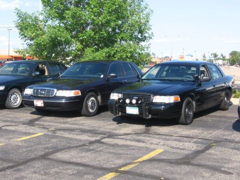 1999 Ford Crown Victoria - Three black Ford Crown Victorias are parked in a lot, one with a push bar and spotlights.