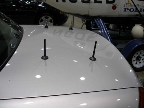 1998 Ford Crown Victoria - Close-up of the silver rear trunk lid of a sedan showing two short black stubby antenna mounts, with a black car and a white police aircraf…