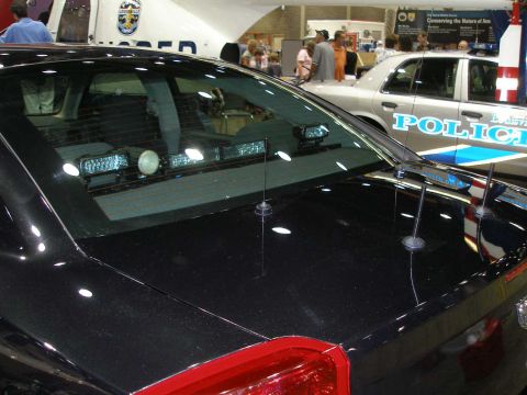 1998 Ford Crown Victoria - Close-up of a black police sedan's rear deck and rear window, showing an interior light bar and dual whip antennas, photographed at an indo…