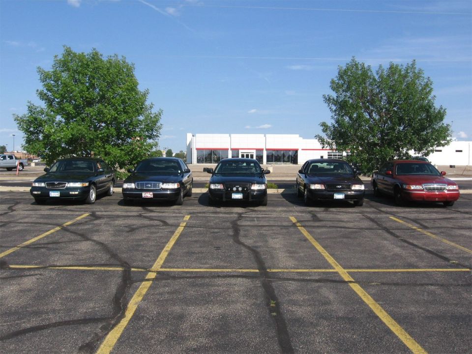 Five Ford Crown Victorias parked in a lot, four are black and appear to be police cars, one is red