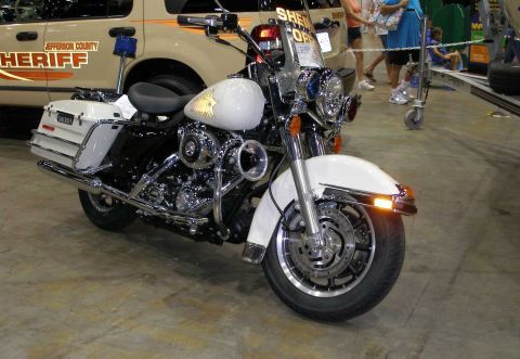 1998 Ford Crown Victoria - A white police touring motorcycle with a gold star emblem on the tank and a 'SHERIFF' saddlebag, displayed at an indoor show in front of a…
