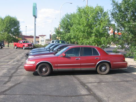 1999 Ford Crown Victoria - A red 1999 Ford Crown Victoria is parked in a lot alongside other vehicles and a red pickup truck.