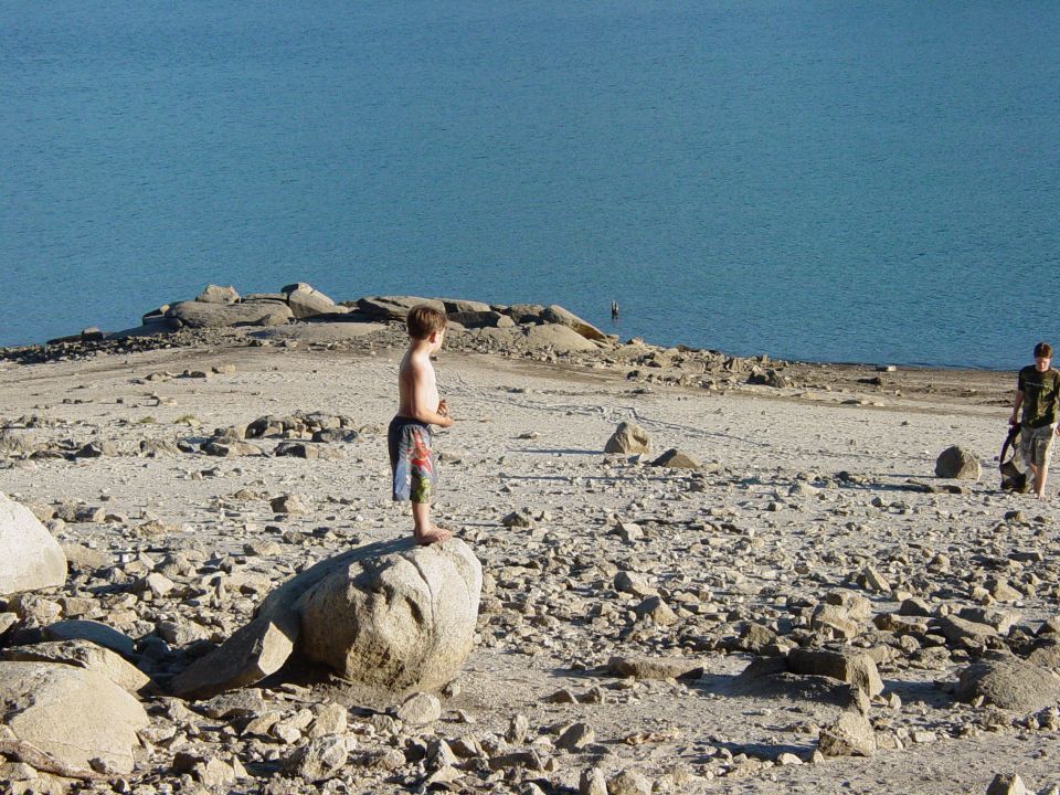 Boy stands on a large rock overlooking a body of water. Another person walks in the distance