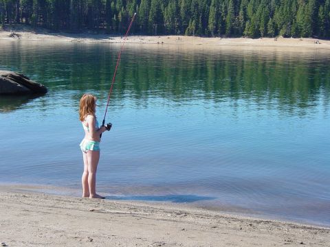 1986 Ford Bronco - A woman stands on a sandy shore, casting a fishing line into a calm lake surrounded by a dense forest.