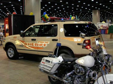 1998 Ford Crown Victoria - A tan Jefferson County Sheriff SUV with orange striping and 'SHERIFF' lettering displayed at an indoor convention hall, with a white Sherif…