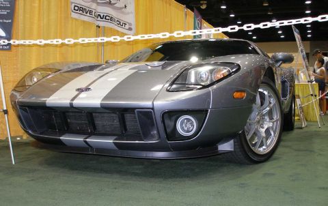 1998 Ford Crown Victoria - A silver-gray Ford GT supercar with dual light-silver racing stripes, shown from a low front three-quarter angle at an indoor car show. The…