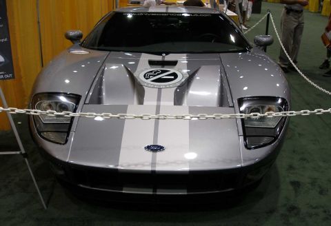 1998 Ford Crown Victoria - A silver Ford GT mid-engine sports coupe with dual dark racing stripes, photographed head-on at an indoor show behind white stanchion chain…