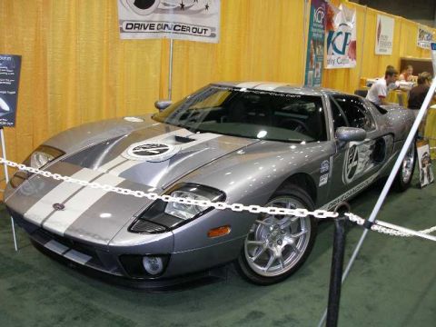 1998 Ford Crown Victoria - A silver Ford GT supercar with dark gray racing stripes displayed on green carpet at an indoor show, roped off with white chains and bearin…