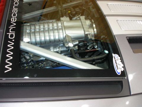 1998 Ford Crown Victoria - View through a rear glass engine cover into the mid-mounted engine bay of a silver sports car, showing a polished intake manifold with prom…