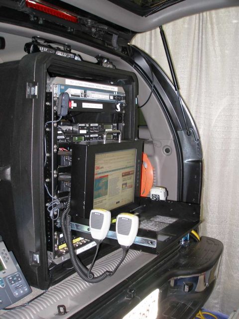 1998 Ford Crown Victoria - A rear cargo area of an SUV outfitted as a mobile command center, with a black equipment rack holding radios, a monitor displaying a CNN.co…