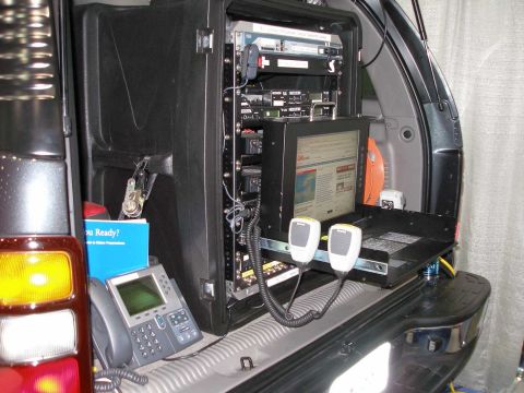 1998 Ford Crown Victoria - The rear cargo area of an SUV outfitted as a mobile command post, featuring a black equipment rack with rackmount radios and electronics, a…