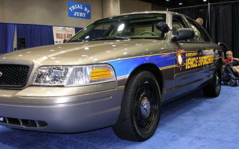 1998 Ford Crown Victoria - A tan/gold Ford Crown Victoria police-style sedan shown from a low front three-quarter angle, marked with blue and yellow striping and 'Ken…