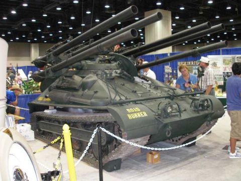 1998 Ford Crown Victoria - A dark olive-green tracked military vehicle marked 'ROAD RUNNER' in yellow, with multiple large recoilless rifle-style barrels angled upwar…