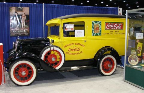 1998 Ford Crown Victoria - Restored 1930s Ford Model A panel delivery truck in yellow over black, displayed indoors at a trade show or exhibit hall. It wears 'Schmidt…