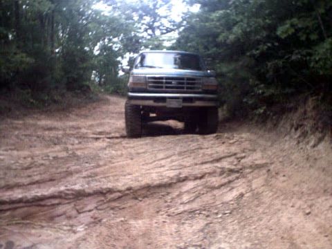 1996 Ford Bronco - A Ford Bronco navigates a muddy, rutted trail surrounded by trees.