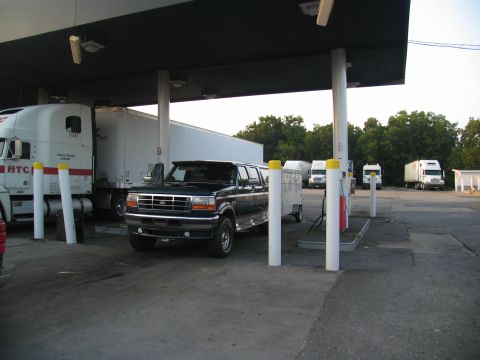 1995 Ford C-350 (Centurion Conversions) - A dark green SUV with a trailer is parked at a gas station, with several semi-trucks visible in the background.