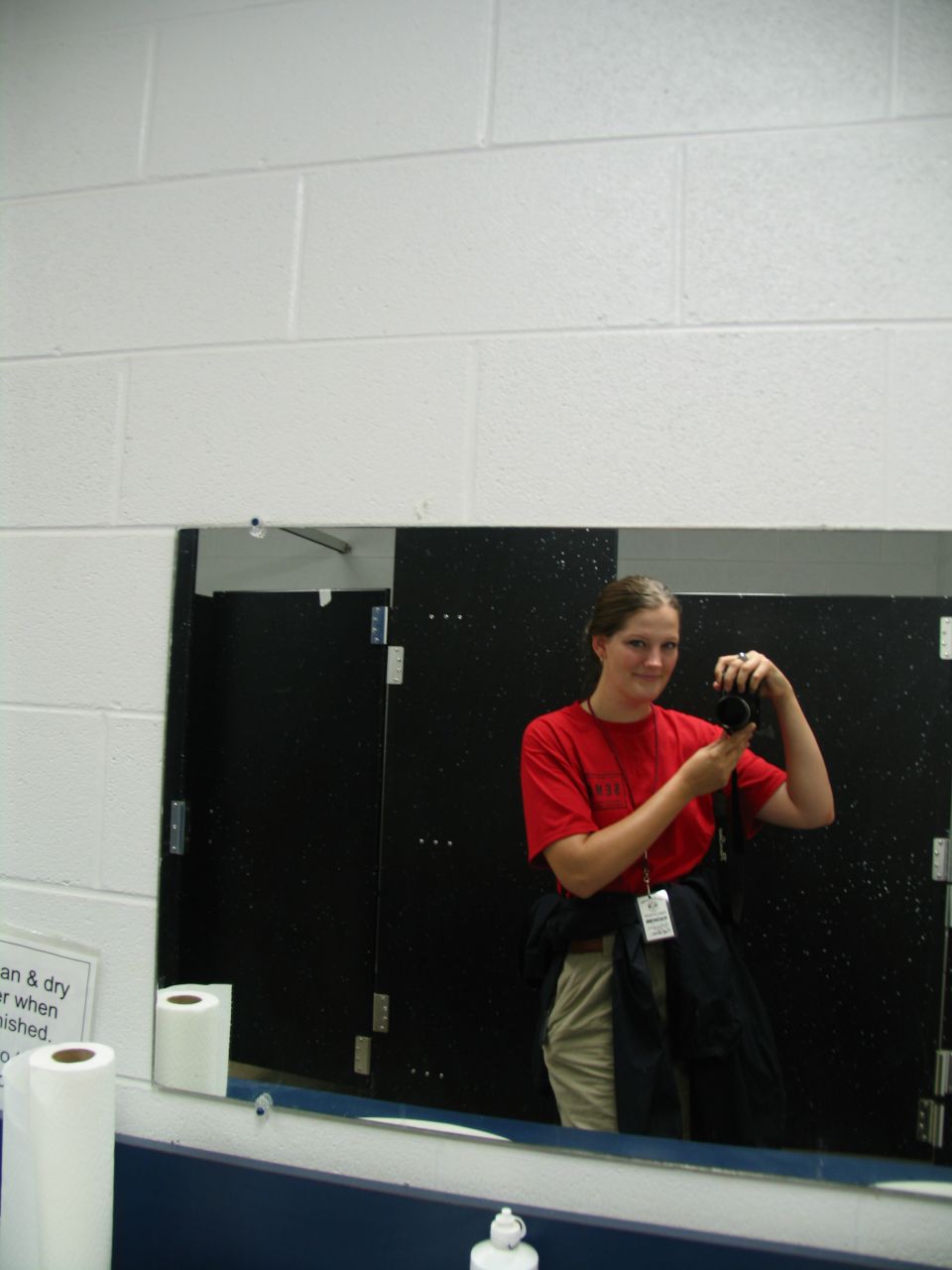 Woman in red shirt and khaki pants takes a mirror selfie in a restroom, holding a camera