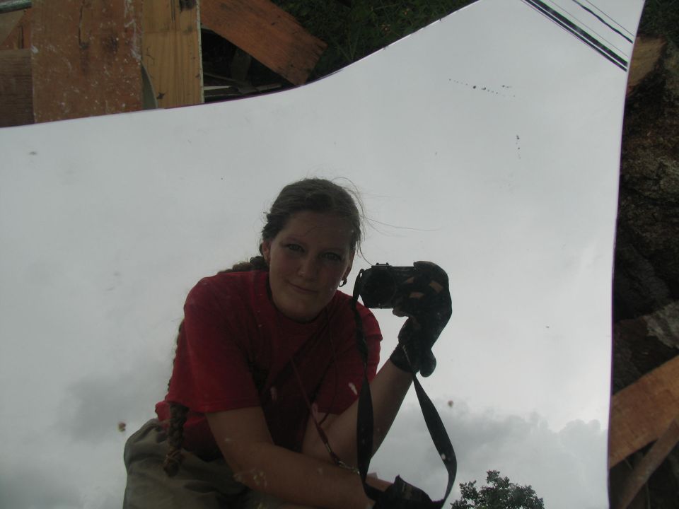 Woman in red shirt and tan shorts holds camera, reflected in a large, flat, white surface with debris around the edges