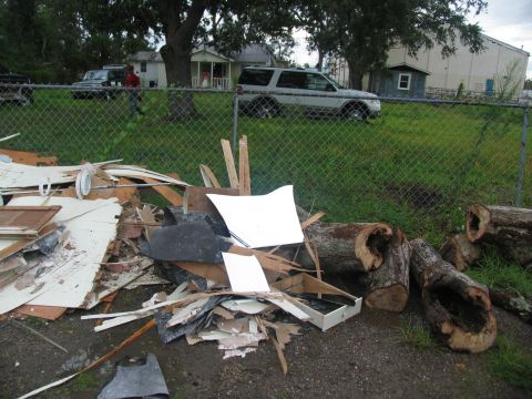 1995 Ford C-350 (Centurion Conversions) - Debris and logs are piled up in front of a fence, with houses and vehicles visible beyond.