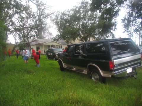 1995 Ford C-350 (Centurion Conversions) - A dark green and silver SUV is parked in a grassy yard near a damaged house, with people in red shirts working in the background.