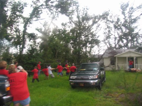 1995 Ford C-350 (Centurion Conversions) - People participate in a tug-of-war game near a dark SUV and a house, possibly after a storm.