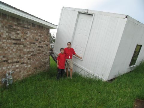 1995 Ford C-350 (Centurion Conversions) - A damaged portable building is tilted on its side next to a brick house after a storm.
